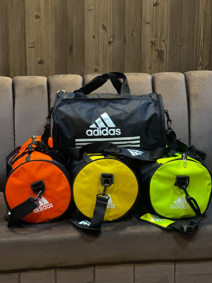 Four Adidas bags in black, orange, yellow, and green on a couch with a wooden wall background.